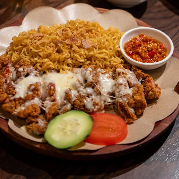 Indonesian fried chicken with melted cheese, noodles, chili sauce, cucumber, and tomato on a wooden plate.